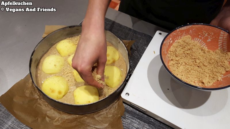 Apfelkuchen mit Streusel in Zubereitung. Über den Boden in den bereits Apfelspalten eingedrückt sind, wird noch vorbereiter Streusel verteilt.
