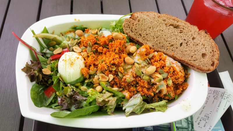 Little Green Rabbit Couscous Salat. In einer Schale mit verschiedenen Blattsalaten, Erdnüssen, Ei und Dressing serviert. Am Rand der Schale liegt eine Scheibe Brot.