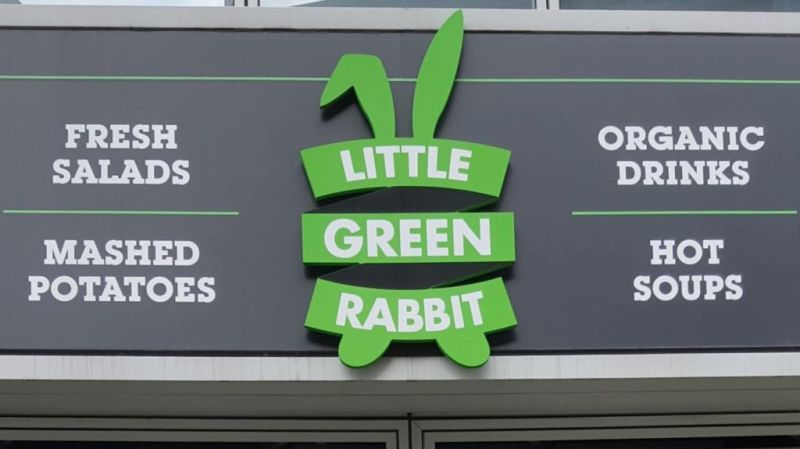 Little Green Rabbit Schild über dem Eingang der Filiale am Checkpoint Charlie. Links steht: Fresh Salads und darunter mashed potatoes, in der Mitte auf einem grünen, stilisierten Hasen mit zwei langen Ohren "Little Green Rabbit" und rechts organic drinks und darunter hot soups.