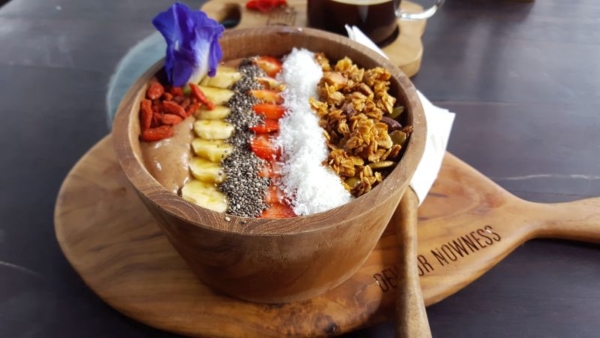 Mudra Café. Macchu Picchu Bowl. Smoothie bowl out of cocoa, banana, date, blueberry, avocado and cashew-milk. Strip-topping out of gojiberries, banana, chia seeds, strawberries, grated coconut and granola. Decorated with a blue flower.