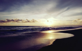 Sonnenuntergang am Strand von Canggu. Es ist schon sehr dunkel, die Sonne spiegelt sich im Ausläufer einer Welle, der Sand erscheint schwarz. Am Horizont ein paar Wolken, die wie ein Schleier über der Sonne liegen.