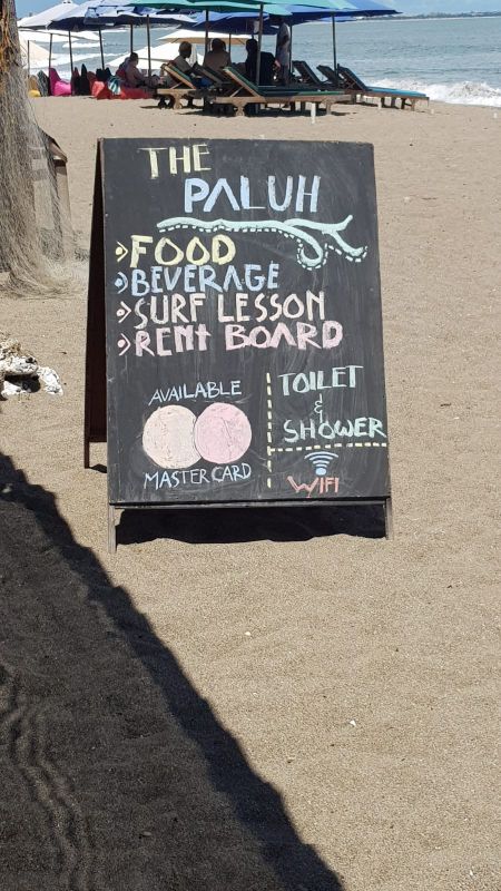 The Paluh Surf and Cafe. Aufsteller im Sand. Darauf, mit bunter Kreide geschrieben, steht: The Paluh. Food. Beverage. Surf Lesson. Rent Board. Available Mastercard und das Mastercard Symbol. Toilet & Shower. WiFi.