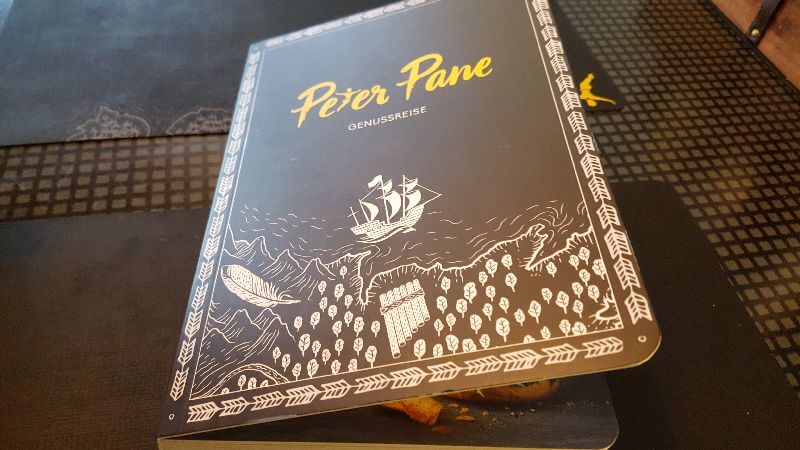 Peter Pane Speisekarte in Buchform. Zu lesen: Peter Pane - Genussreise. Darunter weiß auf schwarz ein gezeichnetes Segelschiff, stilisierte Meeresküste, eine Panflöte, Bäume, Berge und ein Fluss.