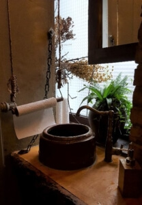 Umami Toilette. Waschbecken rund aus Stein. Patinierter Messingwasserhahn. Auf Seilen hängt eine Papierrolle zum Hände trocknen. Ein paar Pflanzen.